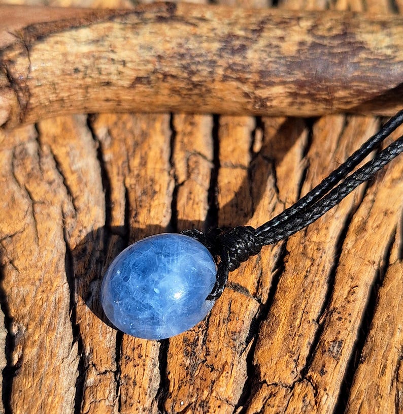 May include: A blue oval-shaped pendant necklace with a black cord. The pendant has a textured surface and is suspended from a braided black cord. The necklace is displayed on a wooden surface.