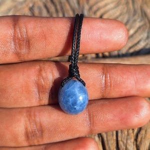 May include: A blue gemstone pendant with a black cord. The gemstone is oval-shaped and has a marbled appearance. The cord is knotted at the top, securing the stone. The pendant is held between fingers, showcasing the jewelry.