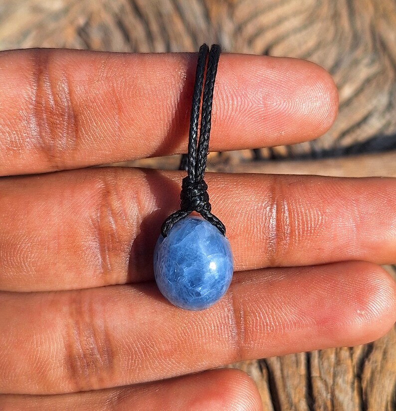 May include: A blue gemstone pendant with a black cord. The gemstone is oval-shaped and has a marbled appearance. The cord is knotted at the top, securing the stone. The pendant is held between fingers, showcasing the jewelry.