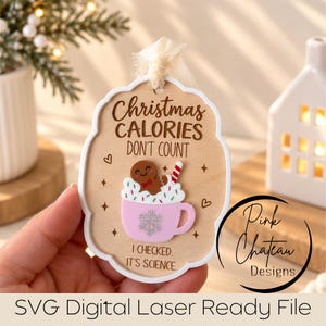 Puede incluir: Un adorno de madera clara con borde blanco y cinta color crema. Incluye el texto "Christmas CALORIES DON'T COUNT I CHECKED. IT'S SCIENCE." y un gráfico en relieve de un hombre de jengibre en una taza rosa con un copo de nieve.