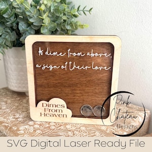 Könnte beinhalten: Holzschild mit einem dunkelbraunen Mittelteil und einem hellen Holzrahmen. Auf dem Schild steht "A dime from above, a sign of their love." Unter dem Text befindet sich ein wolkenförmiger Ausschnitt mit den Worten "Dimes From Heaven". Zwei Münzen sind sichtbar.
