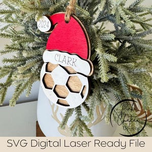 May include: A wooden Christmas bauble shaped like a football with a Santa hat on top. The hat is red with white trim and the football is brown with white stitching. The bauble is personalised with the name "CLARK" and the year "2024".