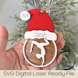 May include: A wooden Christmas bauble shaped like a Santa hat with a gymnast silhouette inside. The bauble is decorated with a woodgrain pattern and has the name "Sarah" and the year "2024" written on it.