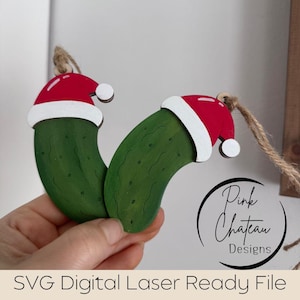 May include: Two green wooden pickle ornaments with black speckles, each wearing a red and white Santa hat, suspended by twine. The text "SVG Digital Laser Ready File" is at the bottom. Festive Christmas decor.