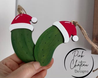 May include: Two wooden pickle ornaments, painted green with black speckles, each wearing a red and white Santa hat. The ornaments are suspended by twine. The text "SVG Digital Laser Ready File" is at the bottom.