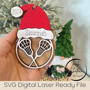 May include: A wooden Christmas bauble featuring a red Santa hat, a lacrosse stick design, and the name "Savannah" inscribed on it. The bauble also has a small tag with the year "2024" on it.