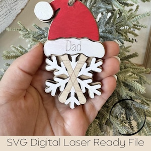 May include: A wooden Christmas decoration featuring a red Santa hat with a white trim and the word "Dad". Below is a snowflake design with crossed skis. The ornament is suspended by a twine loop. The overall design is festive.
