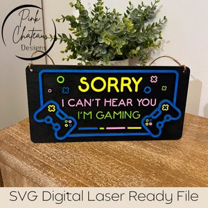 Puede incluir: Un letrero de madera negro con un borde azul neón y texto amarillo que dice "SORRY I CAN'T HEAR YOU I'M GAMING". El letrero presenta dos mandos de juego estilizados en azul y verde con botones blancos.