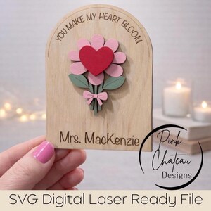 May include: Wooden plaque with a laser-cut flower design. The flower has pink petals, a red heart center, and green leaves. The text "YOU MAKE MY HEART BLOOM" is at the top, and "Mrs. Mackenzie" is at the bottom. The design includes a pink bow.
