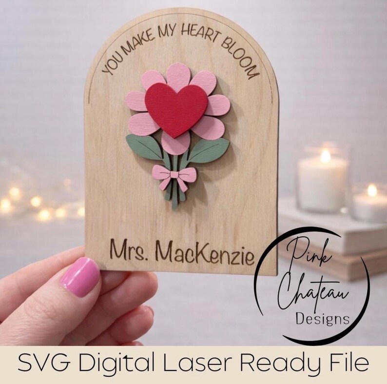 May include: Wooden plaque with a laser-cut flower design. The flower has pink petals, a red heart center, and green leaves. The text "YOU MAKE MY HEART BLOOM" is at the top, and "Mrs. Mackenzie" is at the bottom. The design includes a pink bow.