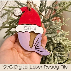 May include: A wooden Christmas ornament shaped like a mermaid wearing a Santa hat. The ornament is painted purple, red, and brown. The ornament has the year 2024 and the name "Sophia" written on it.