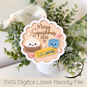 Op de afbeelding: Een geschulpte ronde houten hanger met de tekst Merry Squishmas 2026, een lachende dumpling, een blauw ijsblokje en een geel pakje boter met het label 1 oz NET WT. 28g, versierd met kleine roze hartjes en witte sterren.