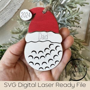 May include: White wooden ornament shaped like a golf ball with a red Santa hat on top. The hat has the word "Dad" written on it and the year "2024" on the side.