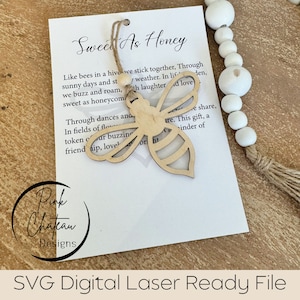 May include: A white card with the text "Sweet as Honey" and a quote about bees. A wooden cutout of a bee is attached to the card with twine. The card is on a wooden surface with a white beaded garland.