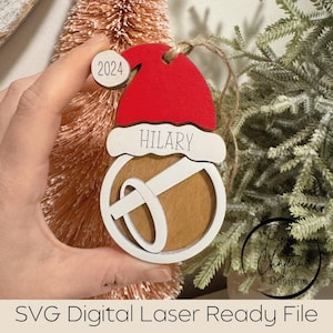 May include: A white wooden Christmas bauble with a red Santa hat, a brown bell, and the name "HILARY" written on it. The ornament also has a small white circle with the number "2024" on it.