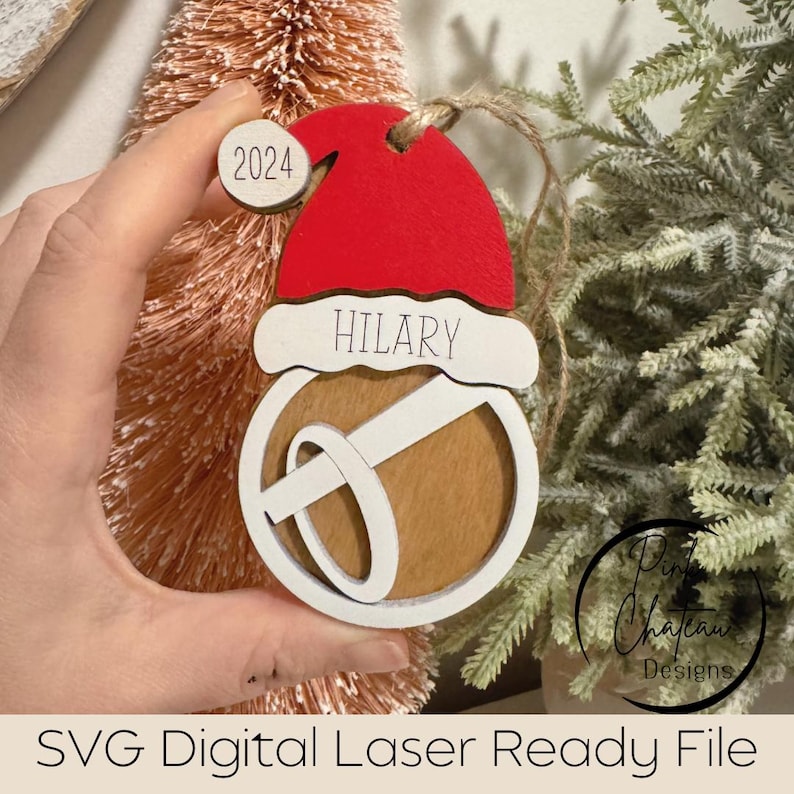 May include: A white wooden Christmas ornament with a red Santa hat, a brown bell, and the name "HILARY" written on it. The ornament also has a small white circle with the number "2024" on it.