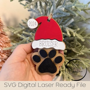 May include: A wooden paw print Christmas ornament with a red Santa hat and the name "BRUISER" on a white banner. The ornament also has the year "2024" on it.