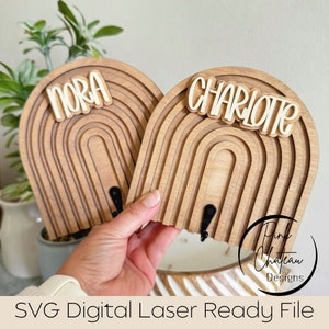 Puede incluir: Dos percheros de pared de madera con forma de arcoíris, uno con la etiqueta "Nora" y el otro "Charlotte". Los ganchos son de madera en capas con un acabado natural y ganchos de metal negro. El texto "SVG Digital Laser Ready File" está en la parte inferior.