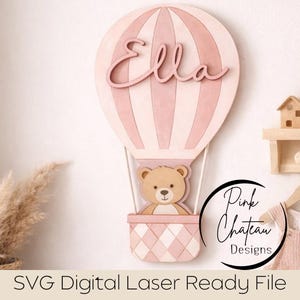 May include: A pink and white wooden hot air balloon wall decor with the name "Ella" in cursive. A teddy bear sits in the basket. The design includes the text "Pink Chateau Designs" and "SVG Digital Laser Ready File".