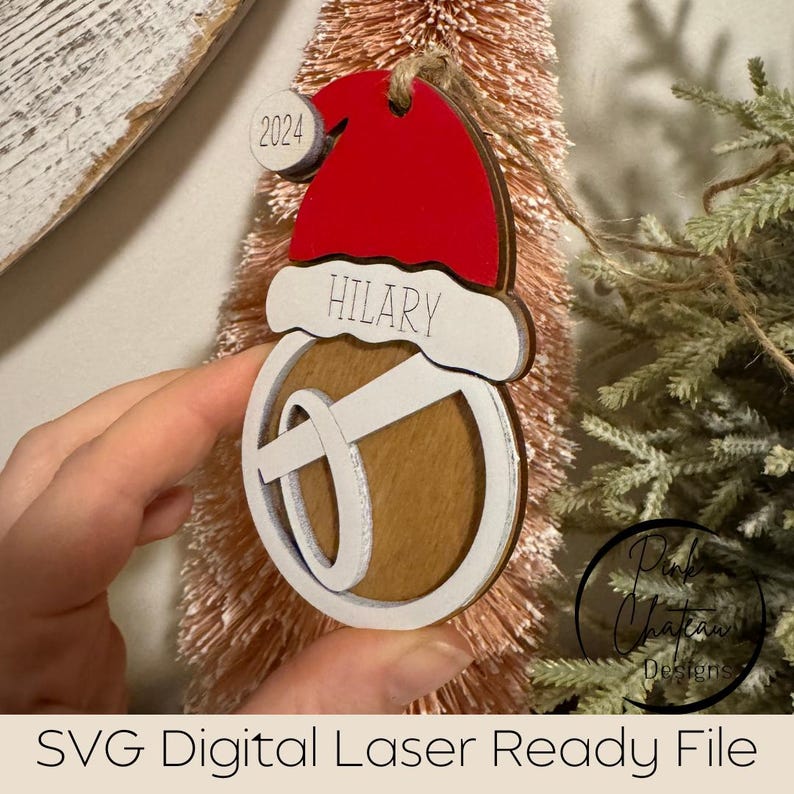 May include: A wooden Christmas ornament shaped like a bell with a Santa hat on top. The ornament is white with a red hat and the name "HILARY" is written on the hat. The year "2024" is written on a small circle on the hat.