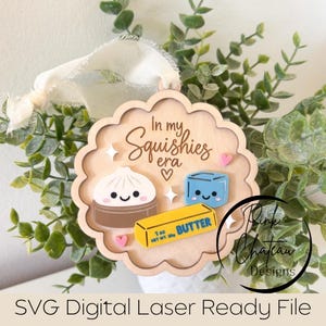 May include: A scalloped wooden ornament featuring the text In my Squishies era, a white dumpling, a blue cube, and a yellow stick of butter labeled 1 oz net wt. 28g. The ornament is topped with a white ribbon and decorated with small pink hearts and white stars.
