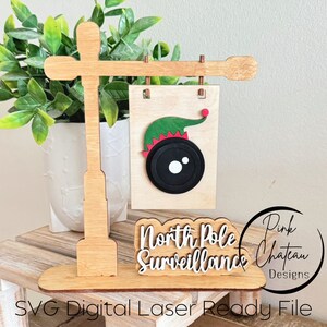 May include: A wooden sign with a camera and elf hat, and the text "North Pole Surveillance". The sign is attached to a wooden pole with a wooden base. The sign is a digital laser-ready file.