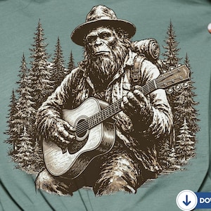 May include: A sage green t-shirt featuring a dark brown graphic illustration of a hairy, bearded creature wearing a hat and carrying a backpack while playing an acoustic guitar. The background of the graphic depicts a dense forest of pine trees.