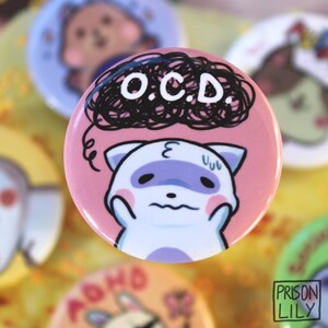 May include: A circular pinback button with a pink background featuring a cartoon ferret with a stressed expression. Above the ferret is a black scribbled cloud with the white text O.C.D. written inside. The button has a glossy finish.