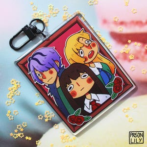 Könnte beinhalten: Ein klarer Acryl-Schlüsselanhänger mit drei Anime-Charakteren in einem roten Rahmen, jeder mit überraschtem Gesichtsausdruck. Der Schlüsselanhänger hat einen schwarzen Verschluss und den Text "PRISON LILY". Sternförmiges Konfetti umgibt den Anhänger.