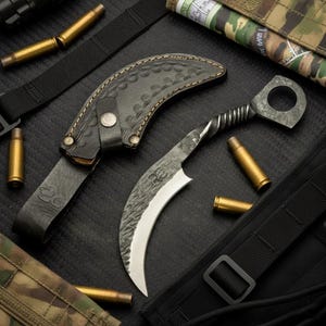 Puede incluir: Un cuchillo karambit curvo con hoja de metal oscuro texturizada, mango retorcido y anillo para el dedo. El cuchillo se acompaña de una funda de cuero negro con costuras decorativas y cierre a presión, rodeado de varios casquillos de bala.