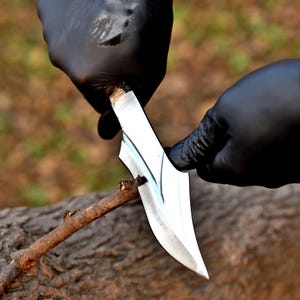 May include: A silver knife with a curved blade is used to carve a small wooden stick. The knife is held by hands wearing black gloves. The background is blurred, showing a natural outdoor setting.