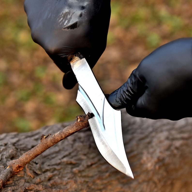 May include: A silver knife with a curved blade is used to carve a small wooden stick. The knife is held by hands wearing black gloves. The background is blurred, showing a natural outdoor setting.