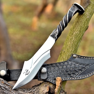 May include: A handcrafted knife with a twisted metal handle and a silver blade. The blade is resting on a black leather sheath with a woven pattern. The knife has a unique design and is likely used for outdoor activities.