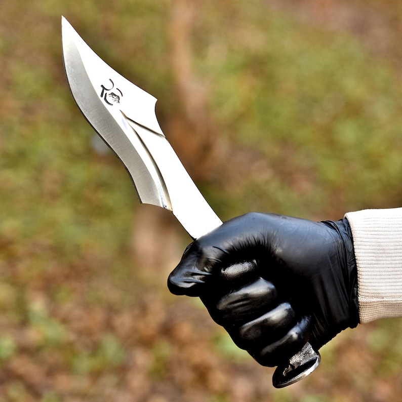 May include: A silver-colored knife with a curved blade and a decorative emblem near the handle. The knife is held by a gloved hand, with a blurred green and brown background.
