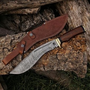 Op de afbeelding: Een Damascus stalen kukri mes met een donker houten handvat en messing accenten, liggend op een stuk hout. Een bruin leren schede bevindt zich achter het mes. Het lemmet heeft een gebogen vorm en een gedetailleerd patroon.