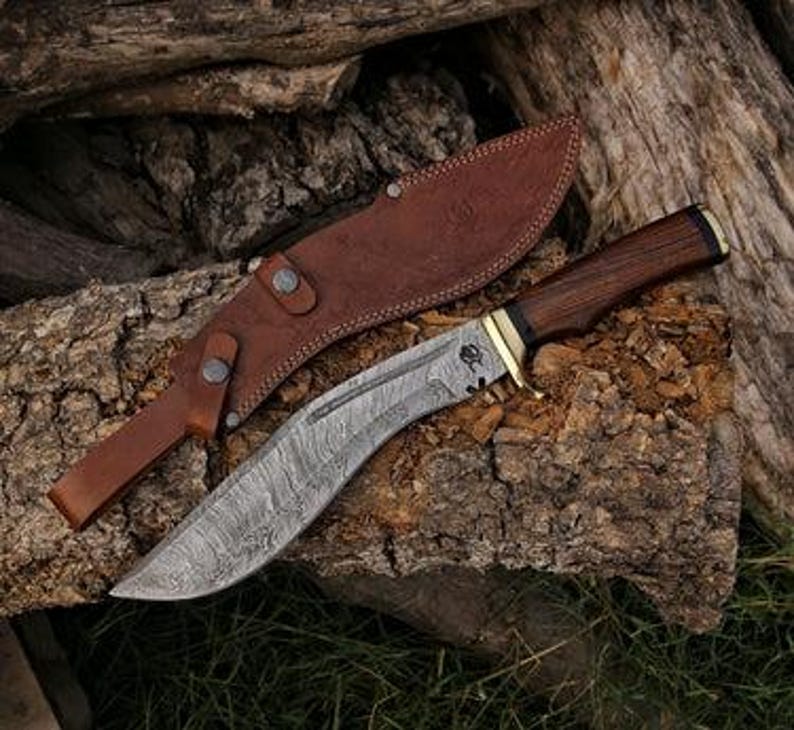 Op de afbeelding: Een Damascus stalen kukri mes met een donker houten handvat en messing accenten, liggend op een stuk hout. Een bruin leren schede bevindt zich achter het mes. Het lemmet heeft een gebogen vorm en een gedetailleerd patroon.
