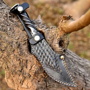 May include: A handcrafted knife with a twisted metal handle and a black leather sheath. The sheath features a woven pattern and is secured with a button. The knife is displayed on a tree branch.