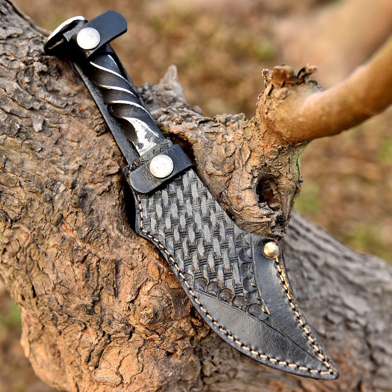 May include: A handcrafted knife with a twisted metal handle and a black leather sheath. The sheath features a woven pattern and is secured with a button. The knife is displayed on a tree branch.