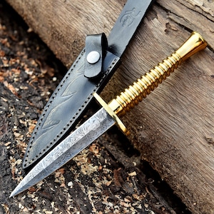 May include: A gold-handled dagger with a patterned blade and a black leather sheath. The dagger is resting on a wooden surface.
