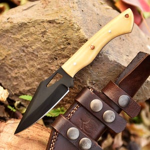 Peut inclure: Un couteau de chasse noir et crème avec un fourreau en cuir. Le couteau a une lame noire et un manche de couleur crème avec un petit trou en haut. Le fourreau est en cuir brun avec trois boutons pression en métal.