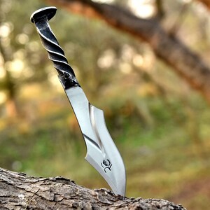 May include: A handcrafted metal knife with a twisted handle and a curved blade. The blade is silver, and the handle is black and silver. The knife is embedded in a piece of wood, showcasing its sharp edge and unique design.
