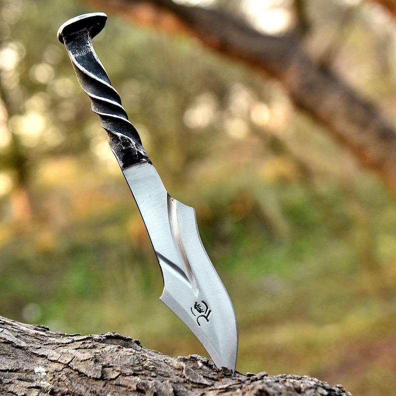May include: A handcrafted metal knife with a twisted handle and a curved blade. The blade is silver, and the handle is black and silver. The knife is embedded in a piece of wood, showcasing its sharp edge and unique design.