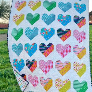 May include: A colorful quilt featuring a pattern of heart shapes in various colors and patterns. The hearts are arranged on a white background. The quilt has a black and white striped border and is displayed outdoors on a grassy surface.