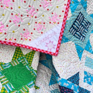 May include: A patchwork quilt featuring a white background with pink and red star patterns, accented by a red and white checkered border. A white corner label displays the text LUMINOVA and a star icon. Various blue and green fish motifs appear.