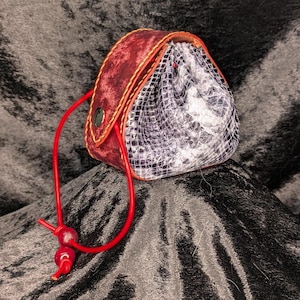 Könnte beinhalten: Ein kleiner, burgunderfarbener Lederbeutel mit Kordelzug. Der Beutel hat ein Schlangenmuster-Innenfutter und eine rote Kordel. Die Kanten sind mit gelbem Faden genäht, und ein silberner Knopf sichert die Klappe.