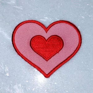 Op de afbeelding: Een roze hartvormige patch met een kleiner rood hart erin. De patch is gemaakt van stof en heeft een gestikte rand.