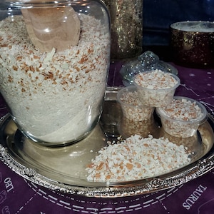 May include: A glass jar filled with crushed eggshells, a wooden scoop, and smaller containers of crushed eggshells on a silver tray. The tray is on a purple surface with a star pattern.