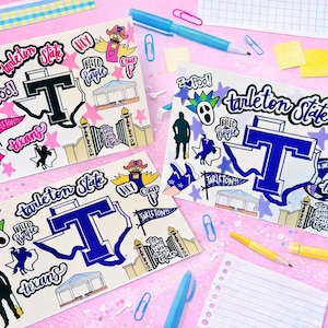 May include: Four Tarleton State University themed stickers with various illustrations and text, including the university's logo, mascot, and phrases like "I Pool!" and "Hey". The stickers are on a pink surface with pens, paperclips, and other stationery.