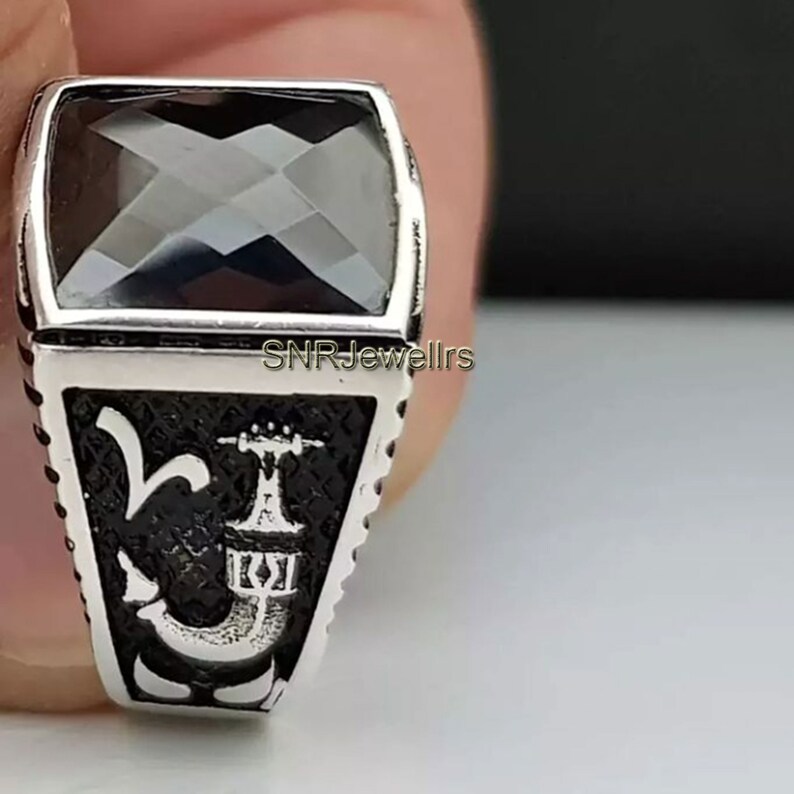 May include: A silver ring with a large, faceted, black gemstone. The ring has a rectangular face with a black background and a decorative design. The text "SNR Jewellrs" is visible on the side of the ring.
