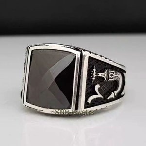May include: A silver ring with a large, square, black gemstone. The ring has a detailed design on the sides, and the text "SNRJewellrs" is visible on the band.